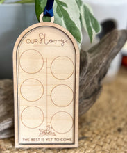 Layered Baltic birch Our Story milestone ornament with six circular coin spaces, engraved holly, the words The Best Is Yet to Come, and navy satin ribbon.