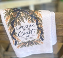 White waffle-weave tea towel with Christmas on the Coast and Outer Banks North Carolina lettering framed by evergreen branches, pinecones, white berries, golden sea oats, and navy ribbon.
