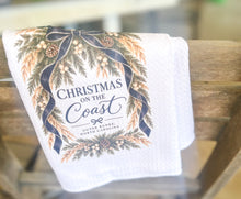 White waffle-weave tea towel with Christmas on the Coast and Outer Banks North Carolina lettering framed by evergreen branches, pinecones, white berries, golden sea oats, and navy ribbon.
