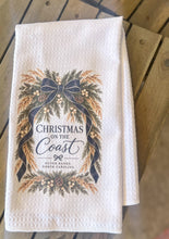 White waffle-weave tea towel with Christmas on the Coast and Outer Banks North Carolina lettering framed by evergreen branches, pinecones, white berries, golden sea oats, and navy ribbon.
