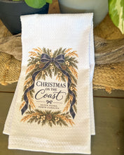 White waffle-weave tea towel with Christmas on the Coast and Outer Banks North Carolina lettering framed by evergreen branches, pinecones, white berries, golden sea oats, and navy ribbon.
