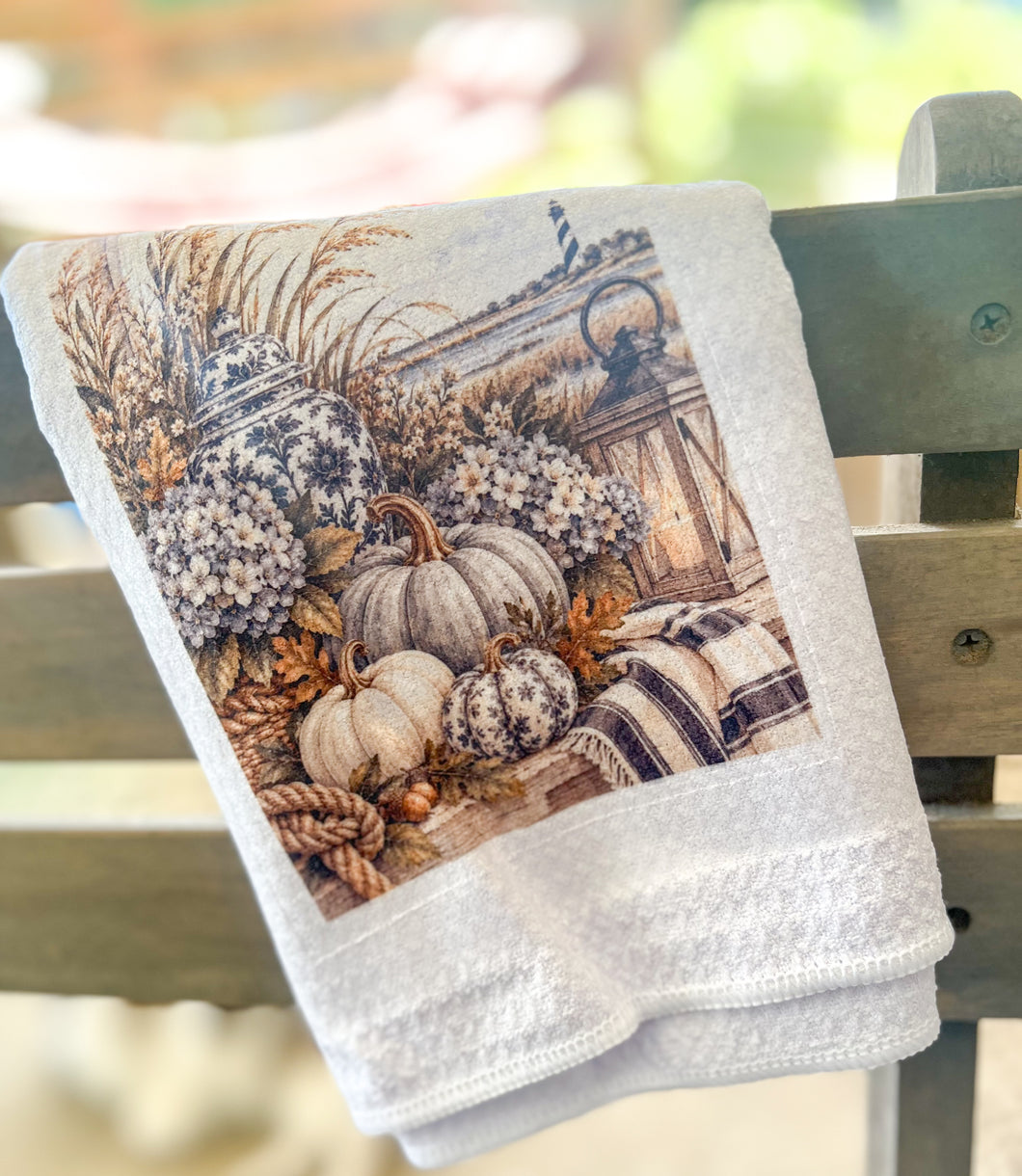 White waffle-weave tea towel printed with cream pumpkins, hydrangeas, navy chinoiserie jars, a lantern, sea oats, rope, plaid fabric, and a distant lighthouse.
