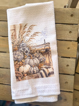 White waffle-weave tea towel printed with cream pumpkins, hydrangeas, navy chinoiserie jars, a lantern, sea oats, rope, plaid fabric, and a distant lighthouse.
