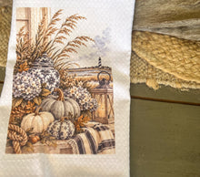 White waffle-weave tea towel printed with cream pumpkins, hydrangeas, navy chinoiserie jars, a lantern, sea oats, rope, plaid fabric, and a distant lighthouse.
