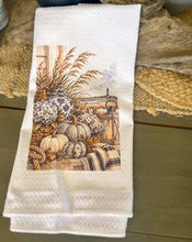 White waffle-weave tea towel printed with cream pumpkins, hydrangeas, navy chinoiserie jars, a lantern, sea oats, rope, plaid fabric, and a distant lighthouse.
