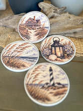 Four round ceramic coasters featuring an autumn coastal cottage, lantern with hydrangeas and shells, quiet shoreline with sea oats, and black-and-white lighthouse.
