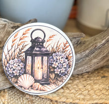 Four round ceramic coasters featuring an autumn coastal cottage, lantern with hydrangeas and shells, quiet shoreline with sea oats, and black-and-white lighthouse.
